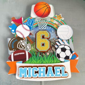 May include: A colorful sports-themed cake topper featuring a basketball, baseball, football, soccer ball, and volleyball. The number 6 is in gold glitter, and the name "MICHAEL" is displayed on a banner. Includes a basketball hoop and stars.