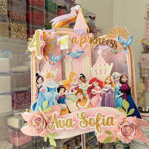 May include: A pink and gold princess-themed cake topper featuring a castle, the number 4, and the text "Ava Sofia." The topper includes images of various Disney princesses and decorative flowers.