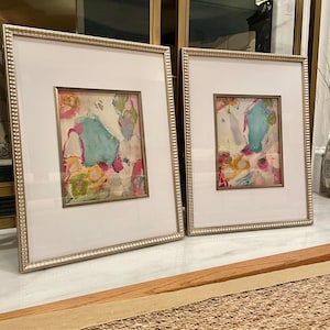 May include: Two framed abstract paintings with silver frames. The paintings are on white backgrounds with colorful abstract designs. The paintings are displayed on a wooden surface with a woven rug in the foreground.