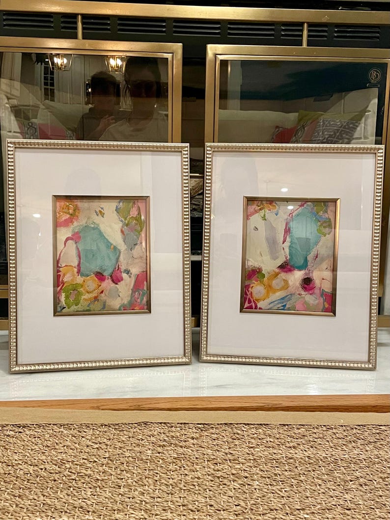 May include: Two framed abstract paintings with gold frames and white mats. The paintings are both colorful and feature a mix of pink, blue, yellow, and green colors. The paintings are displayed on a light brown surface.