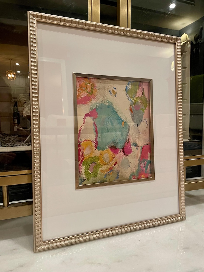 May include: A framed abstract painting with a white mat and a gold frame. The painting features a variety of colors including pink, blue, green, and yellow. The painting is abstract and features a variety of shapes and textures.