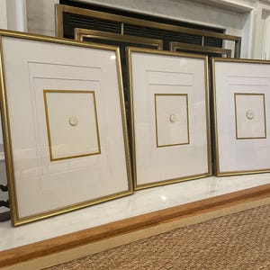 Puede incluir: Tres obras de arte enmarcadas con marcos dorados y paspartús blancos. Cada marco presenta un borde interior dorado y un pequeño diseño circular en el centro. Los marcos se exhiben sobre una superficie blanca.