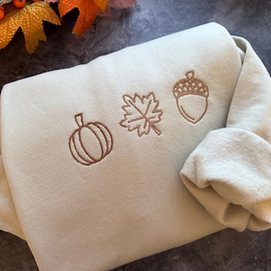 May include: A light green sweatshirt with brown embroidered autumn-themed designs. The designs include a pumpkin, a leaf, and an acorn.