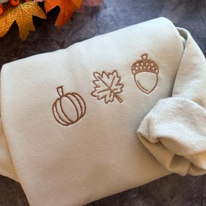 May include: A light green sweatshirt with brown embroidered autumn-themed designs. The designs include a pumpkin, a leaf, and an acorn.