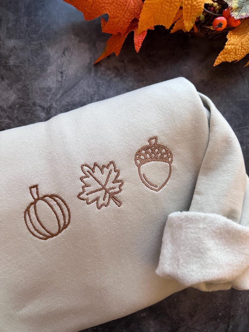 May include: A light green sweatshirt with brown embroidered autumn-themed designs including a pumpkin, a leaf, and an acorn.
