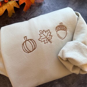 May include: A light green sweatshirt with brown embroidered autumn-themed designs. The designs include a pumpkin, a leaf, and an acorn.