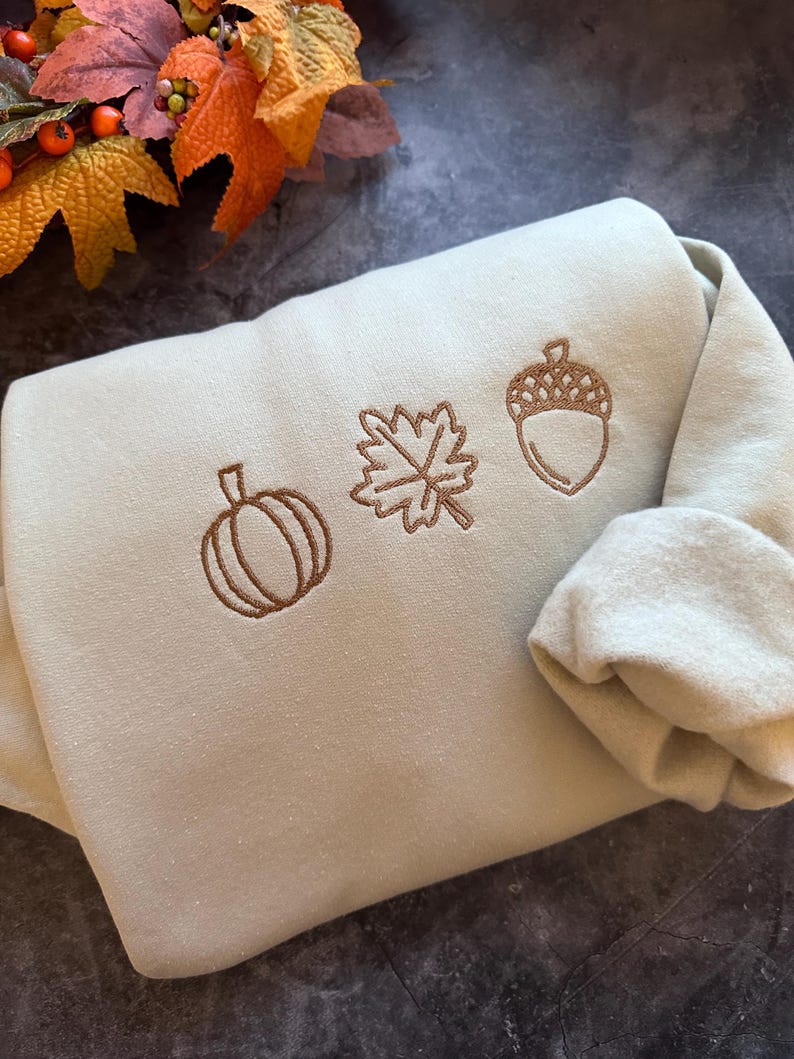 May include: A light green sweatshirt with brown embroidered autumn-themed designs. The designs include a pumpkin, a leaf, and an acorn.