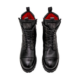 May include: Pair of black leather combat boots with a textured finish. The boots feature a lace-up design, side zippers, and a red interior lining. The boots have a lug sole and a logo on the tongue.