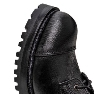 May include: Close-up of a black leather boot. The boot features a textured leather upper, a thick, lugged sole, and visible stitching details. The laces are partially visible. The overall design suggests a durable, work-style boot.