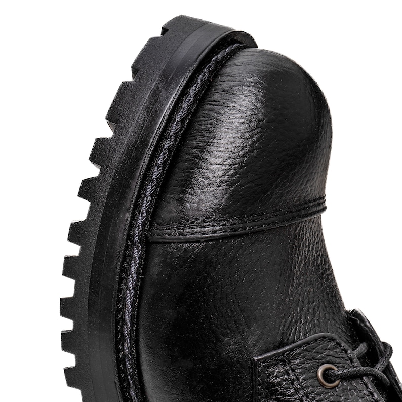 May include: Close-up of a black leather boot. The boot features a textured leather upper, a thick, lugged sole, and visible stitching details. The laces are partially visible. The overall design suggests a durable, work-style boot.