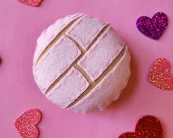 May include: A felt concha pastry with a pink and tan design, surrounded by glittery heart-shaped decorations in shades of pink, red, and purple, set against a pink background.