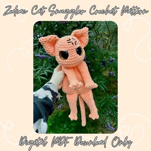 Puede incluir: Un juguete de amigurumi de gato tejido a crochet en color melocotón con ojos negros. El gato tiene una expresión gruñona y está siendo sostenido por una mano. El fondo es una imagen borrosa de follaje verde.