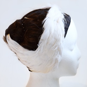 May include: A white feather headband with a white feather accent on the side. The headband is worn on a white mannequin head with black hair.