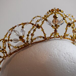 May include: A handcrafted tiara with a delicate design. The crown features a network of gold-colored wire adorned with clear and amber-colored beads. The tiara is displayed on a white, rounded form, showcasing its intricate details.