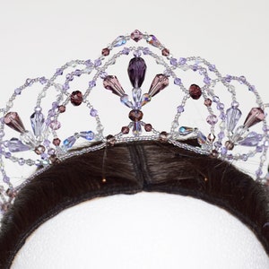 May include: A delicate, beaded tiara with a floral design. The tiara is made with clear, iridescent, and purple crystals. It is worn on a mannequin head with dark brown hair.