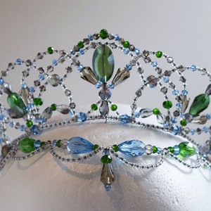 May include: A silver wire tiara with blue, green, and clear crystals. The tiara is decorated with a delicate floral design.