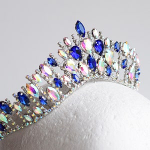 May include: A silver tiara with blue and iridescent rhinestones. The tiara is designed in a cascading pattern with a pointed top.