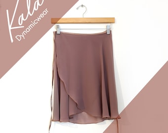 Può includere: Una gonna a portafoglio color rosa polvere realizzata in tessuto leggero e trasparente, appesa a una gruccia di legno. La gonna presenta una chiusura a laccio sul lato e un orlo curvo. Il testo Kala Dynamicwear è visibile in alto a sinistra.