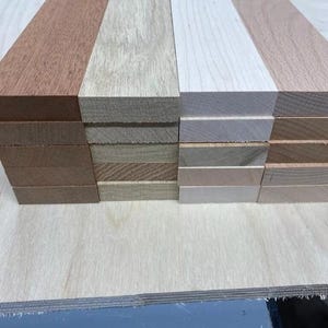 Puede incluir: Cuatro pilas de tablones de madera en diferentes colores. La pila de la izquierda es de color marrón rojizo, la siguiente pila es de color marrón claro con una veta más oscura, la tercera pila es de color blanco con una veta marrón claro y la última pila es de color marrón claro con una veta más oscura.