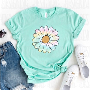 K&ouml;nnte beinhalten: Ein hellgr&uuml;nes T-Shirt mit einem Batik-G&auml;nsebl&uuml;mchen-Design. Die Blume hat eine gelbe Mitte und regenbogenfarbene Bl&uuml;tenbl&auml;tter.