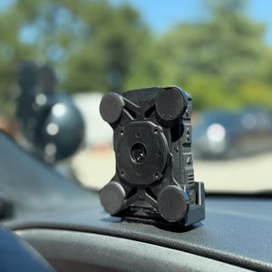 May include: A black, rectangular car phone mount with four circular suction cups. The mount is attached to a car dashboard. The background is blurred, showing trees and a blue sky.