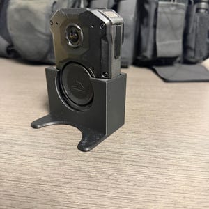 May include: A black body camera with a lens and a speaker, mounted on a black stand. The camera is rectangular with rounded edges. The stand has a curved base and a circular opening for the speaker. The background includes a black tactical vest.