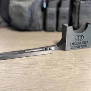May include: A black 3D printed plastic accessory featuring a long, narrow arm with a slotted track and a wider, rectangular base section. The base displays the embossed text DEBIASIO DESIGNS with a logo above it and a curved cutout on the top edge.