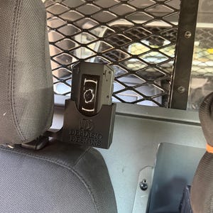 May include: A black plastic holder for a device, mounted on a vehicle headrest. The holder has the text "DEBIASIO DESIGNS" printed on it. The device is black with a white graphic.
