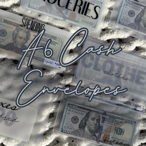 May include: A6 cash envelopes with labels for Groceries, Rent, Spending, Clothes, and Taxes. Each clear envelope contains cash, with the amounts visible. The envelopes are arranged on a white, textured surface.