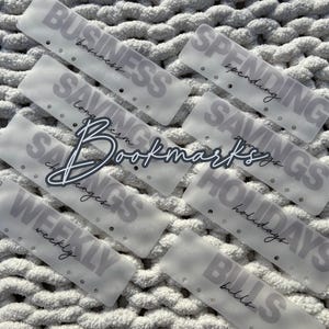 May include: A collection of translucent bookmarks with various financial categories printed on them, including "Business," "Spending," and "Savings." The word "Bookmarks" is written in cursive. The bookmarks are arranged on a textured, light gray surface.