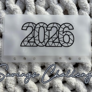 May include: A clear plastic savings envelope with the year "2026" in a geometric black font. The words "Savings Challenge" are written in cursive below the envelope. The background is a white, chunky knit fabric.