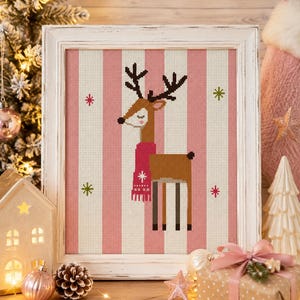 May include: A framed cross-stitch artwork featuring a brown deer with dark antlers wearing a pink fringed scarf. The deer is centred against a background of vertical pink and white stripes, accented with small green and pink star-shaped motifs.