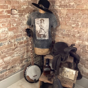 May include: A black and white photo of a young girl holding a rifle, printed on a gray t-shirt. The t-shirt is displayed on a mannequin with a cowboy hat, banjo, saddle, and a bottle of whiskey.