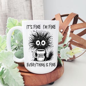 May include: White ceramic mug featuring a black and white illustration of a cartoon cat holding a coffee mug. The text on the mug reads "It's fine I'm fine Everything is fine".