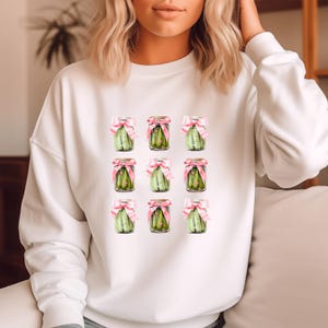 Peut inclure: Sweat-shirt blanc avec un motif de neuf bocaux de cornichons liés avec des rubans roses.