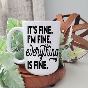 May include: White ceramic coffee mug with black text that reads "It's fine. I'm fine. everything is fine."