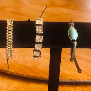 May include: Five different bracelets displayed on a black stand. The bracelets are made of leather, beads, and metal. One bracelet features a turquoise stone, another has a clear crystal, and another has a silver circle with small stones.