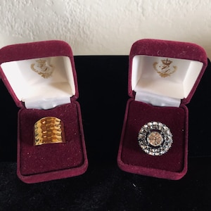 May include: Two ring boxes with velvet interiors. The box on the left contains a gold ring with a textured surface. The box on the right contains a silver ring with a large clear stone surrounded by smaller stones.