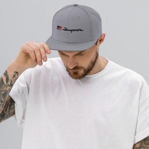 May include: A gray and black baseball cap with the word "Supra" embroidered in black and the letters "GR" in red and black. The cap has a flat brim and a curved crown.