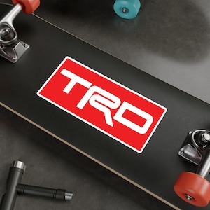 Può includere: Un adesivo rettangolare rosso con il logo TRD bianco. L'adesivo è su uno skateboard nero. Lo skateboard ha ruote rosse e blu. L'immagine è ripresa da un'angolazione leggermente elevata.