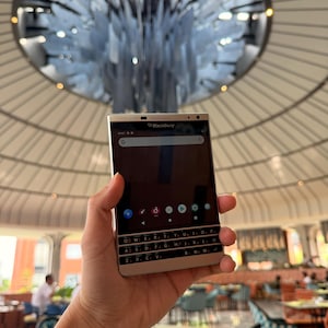 May include: A silver BlackBerry smartphone featuring a physical QWERTY keyboard below a touchscreen display. The screen shows a search bar, app icons, and the BlackBerry logo at the top. The device has a metallic frame and a rectangular shape.