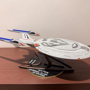 May include: A model of the Sovereign Class Enterprise NCC-1701-E starship, featuring a white and gray saucer section, two long nacelles with blue and red accents, and a black triangular display stand with the text AD ASTRA PER ASPERA.
