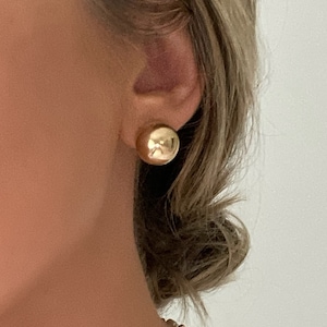 May include: A pair of gold-toned round stud earrings. The earrings are worn by a person with a pearl necklace.
