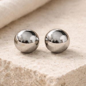 May include: Two shiny, silver-colored, spherical objects. The spheres are reflective, with a smooth surface. They are resting on a light-colored, textured surface. The objects appear to be jewelry, possibly earrings or decorative elements.