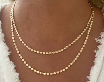 May include: A double-layered gold necklace featuring two delicate chains composed of small, flat, circular metallic discs. The chains are arranged in a tiered style, resting against the neck, with each link reflecting light to create a shimmering effect.
