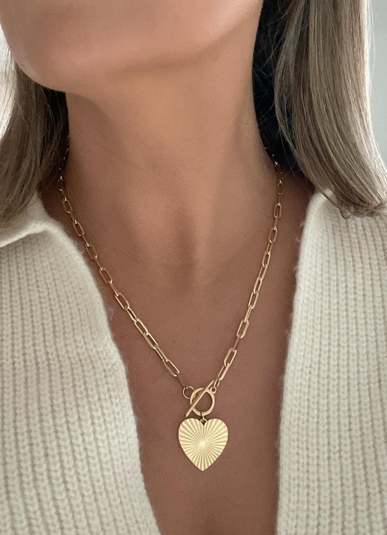 K&ouml;nnte beinhalten: Goldfarbene Halskette mit einem Herzanh&auml;nger mit strukturiertem Sonnenstrahl-Design. Der Anh&auml;nger h&auml;ngt an einer Kette mit einem Knebelverschluss. Die Halskette wird &uuml;ber einem cremefarbenen Pullover getragen.