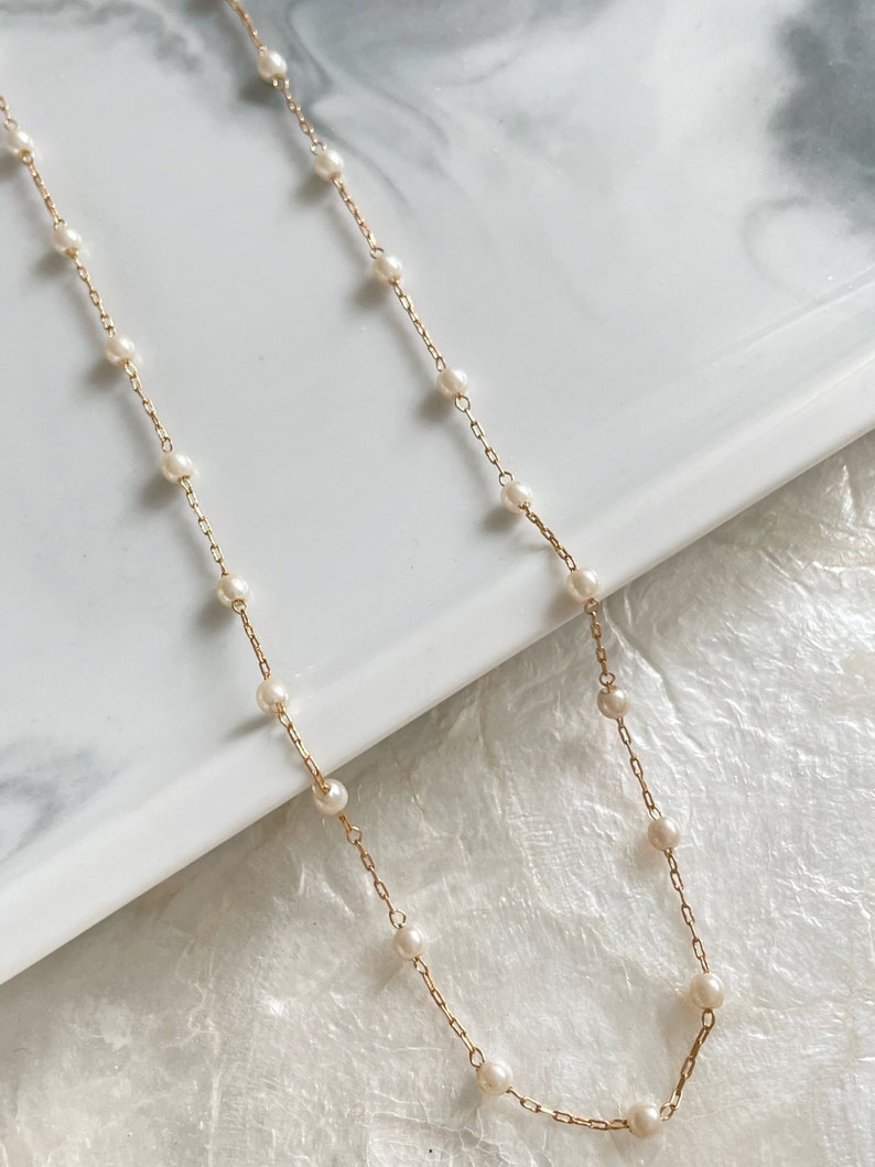May include: A delicate gold chain necklace featuring evenly spaced, small white pearls integrated along the length of the metal links. The necklace is composed of a thin, polished gold-tone chain with round, lustrous white pearls attached at regular intervals.