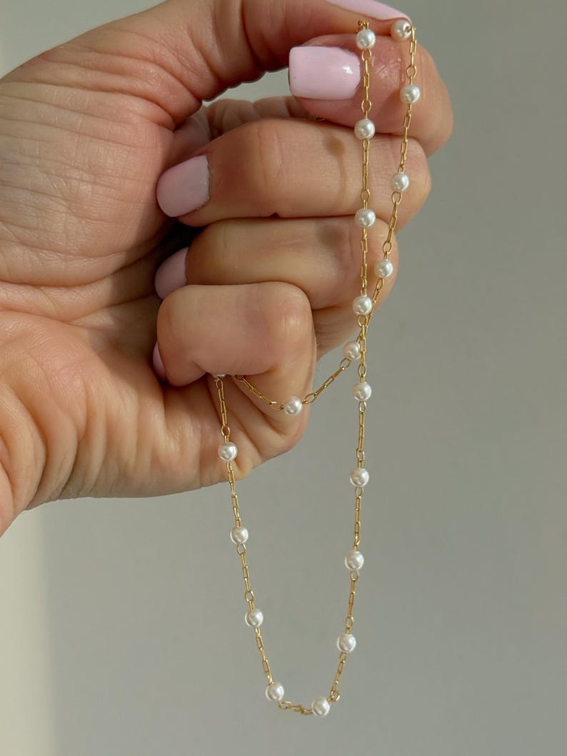 May include: A delicate gold chain necklace featuring evenly spaced small white pearls along the length of the links. The necklace is constructed with a thin metallic gold chain and round, smooth white pearls that are integrated into the chain design.