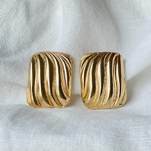 May include: A pair of gold-colored square earrings. Each earring features a ridged, wave-like design. The earrings are displayed on a white, textured surface.