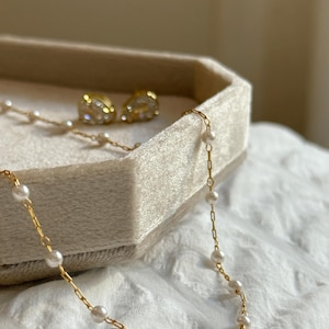 May include: A gold chain necklace featuring small white pearls spaced evenly along the links, accompanied by a pair of gold stud earrings with clear, faceted stones. The jewellery pieces are displayed together in a light beige velvet tray.